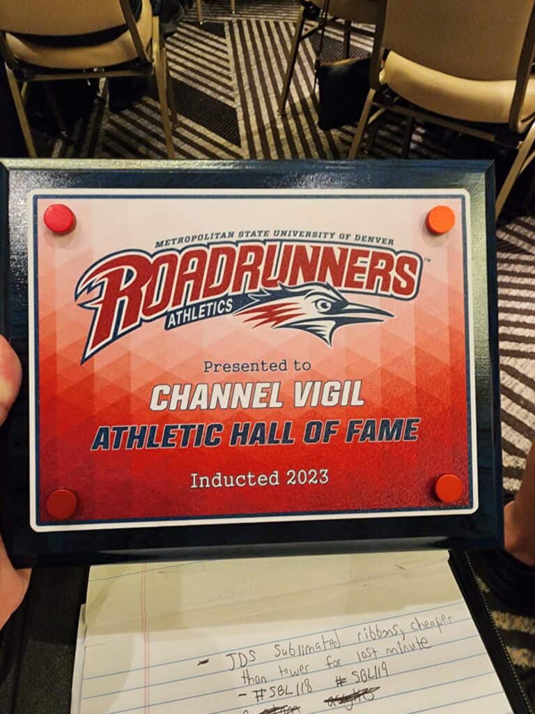 A close-up of a recognition plaque from Metropolitan State University of Denver, featuring the Roadrunners Athletics logo, presented to an individual named Channel Vigil for the Athletic Hall of Fame induction in 2023. The plaque is held over an open notebook with handwritten notes.