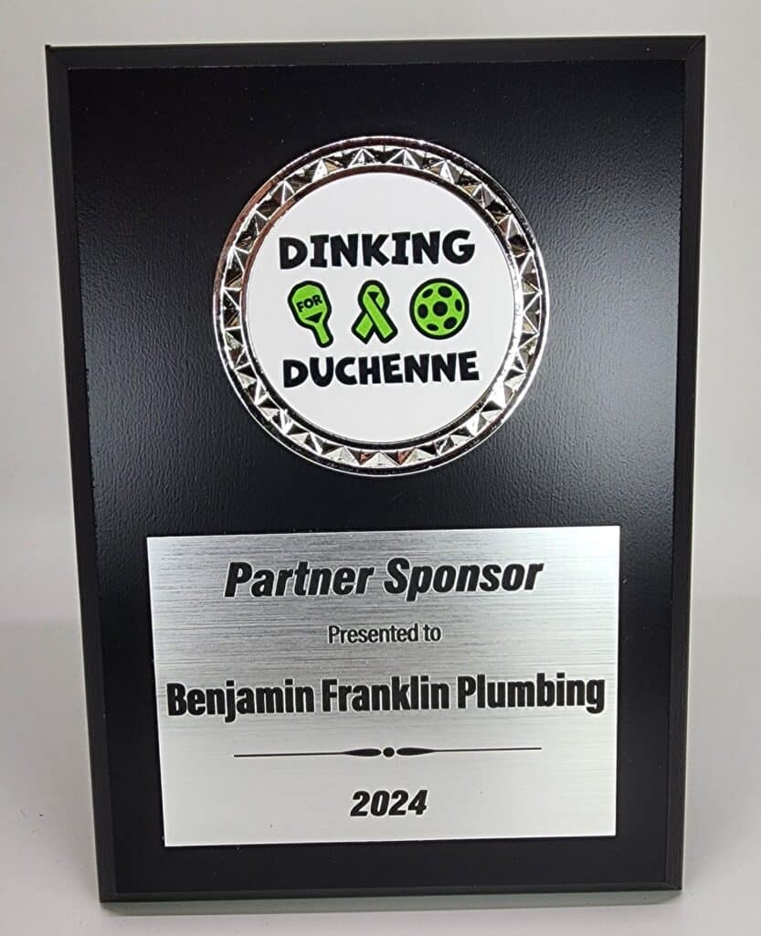 A black plaque featuring a circular emblem at the top with the text &#039;Dinking for Duchenne&#039; and green symbols, followed by a brushed metal plate reading &#039;Partner Sponsor Presented to Benjamin Franklin Plumbing 2024.&#039;