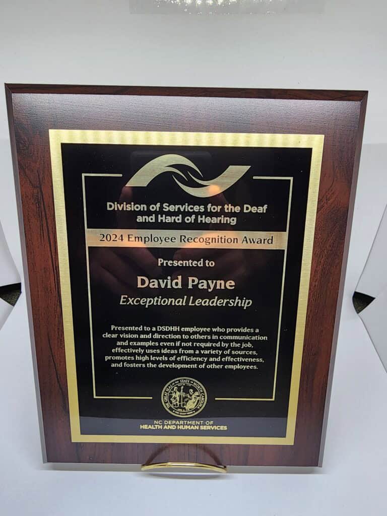 A wooden plaque with a gold and black design, awarded by the Division of Services for the Deaf and Hard of Hearing. It reads &quot;2024 Employee Recognition Award, Presented to David Payne, Exceptional Leadership.&quot; The text highlights leadership qualities and is issued by the NC Department of Health and Human Services.