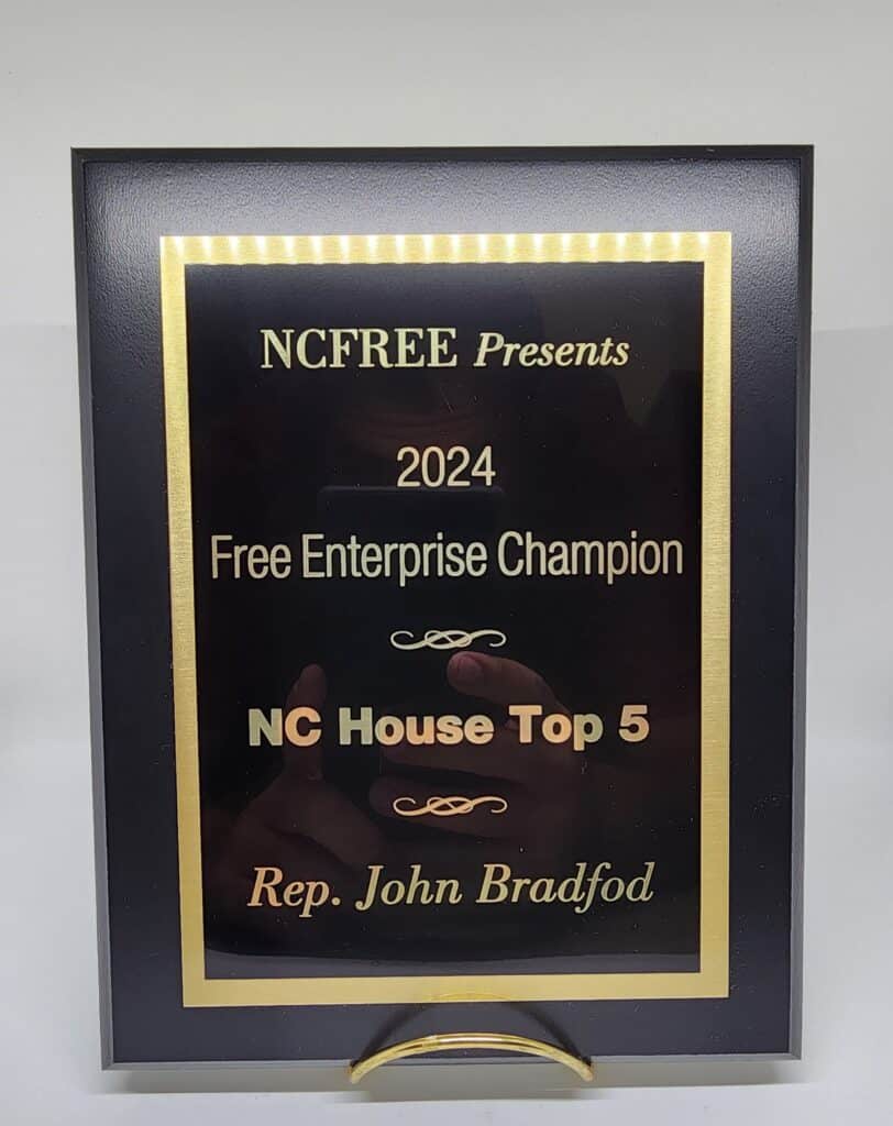 A plaque labeled &quot;NCFREE Presents 2024 Free Enterprise Champion NC House Top 5 Rep. John Bradford,&quot; with a gold and black design.