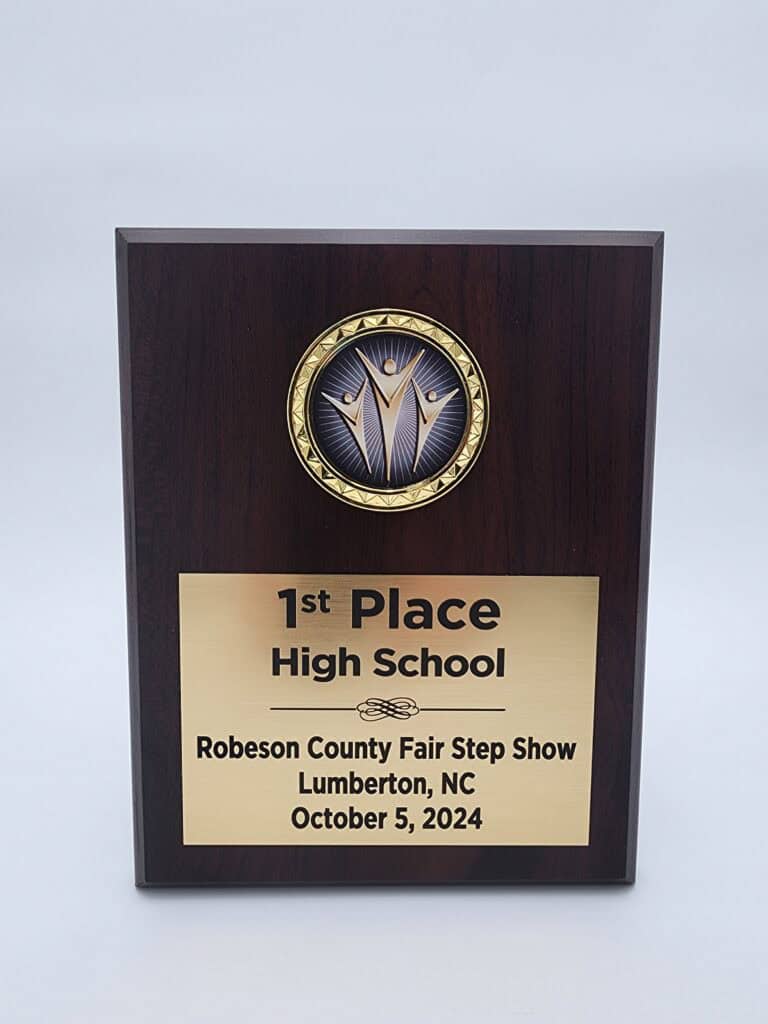 A dark wooden plaque with a circular emblem at the top, featuring three stylized figures with raised arms. Below the emblem, a gold plate is engraved with the text: &quot;1st Place High School, Robeson County Fair Step Show, Lumberton, NC, October 5, 2024.&quot;