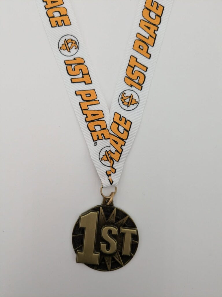 A gold medal with the text '1st' on a circular design, attached to a white ribbon that reads '1st Place' with basketball graphics.