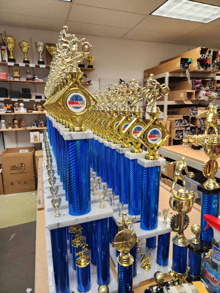 A row of sports trophies with blue columns and gold figures on top, featuring motorcyclist, basketball player, and other athletic figures, displayed in a shop with various other trophies in the background.