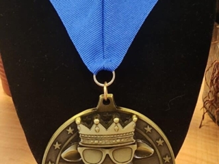A custom medal featuring a goat wearing a crown and sunglasses, with the word 'GOAT' written in large letters below. The medal is attached to a blue ribbon displayed on a black stand.
