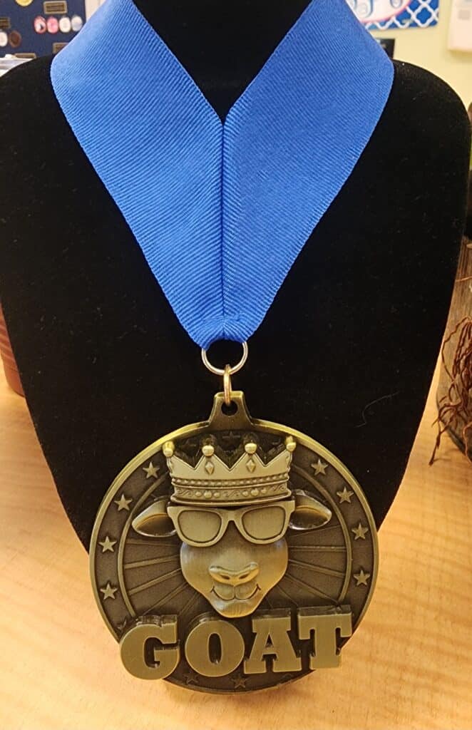 A custom medal featuring a goat wearing a crown and sunglasses, with the word 'GOAT' written in large letters below. The medal is attached to a blue ribbon displayed on a black stand.