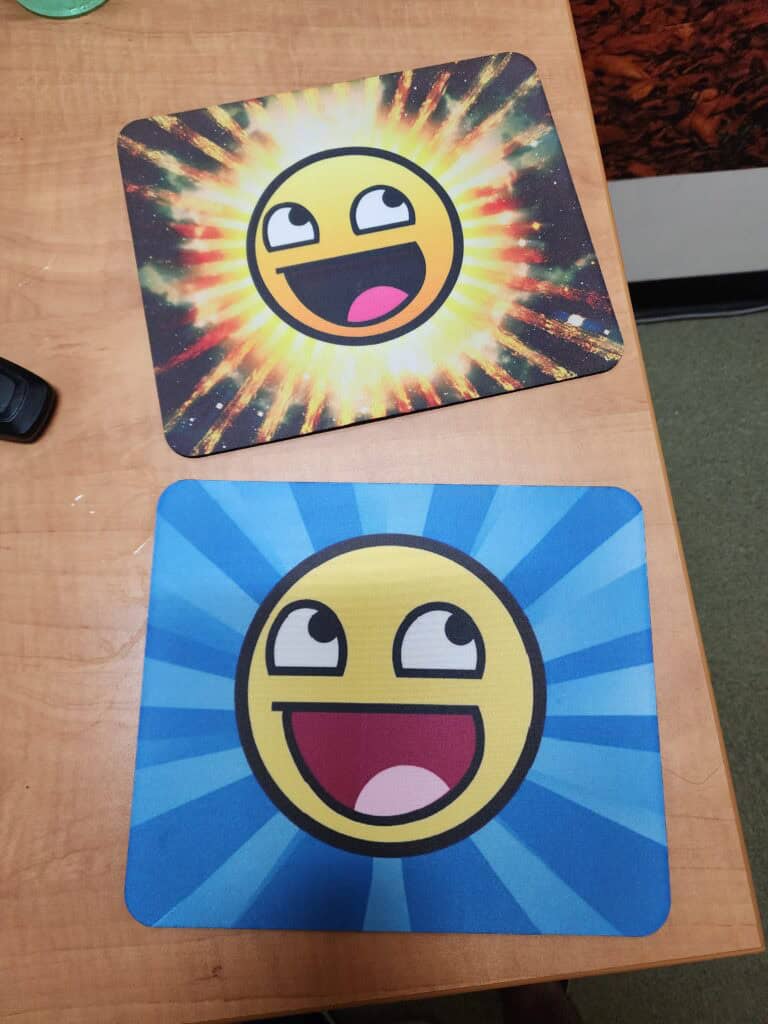 Two rectangular mouse pads on a wooden table, each featuring a yellow cartoon face with a wide-open mouth and eyes looking upwards. The top mouse pad has a colorful explosion background, while the bottom one has a blue radial pattern background.
