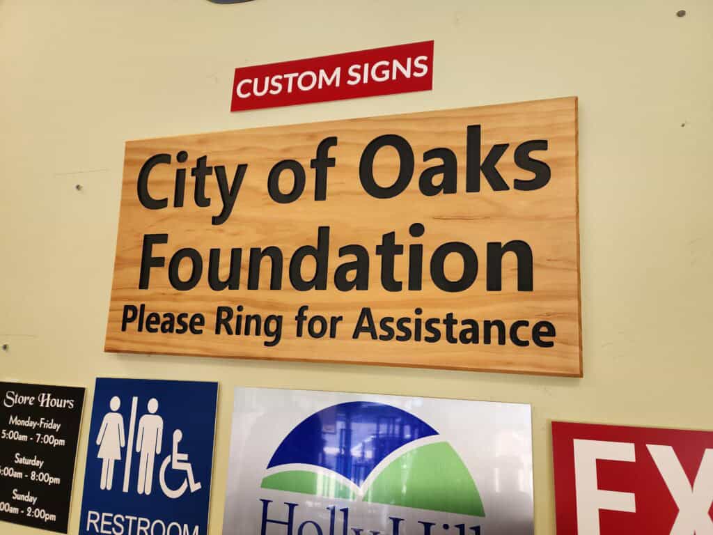 A wooden sign with black text reads "City of Oaks Foundation Please Ring for Assistance," accompanied by additional signs for custom signs, store hours, restrooms, and an exit.