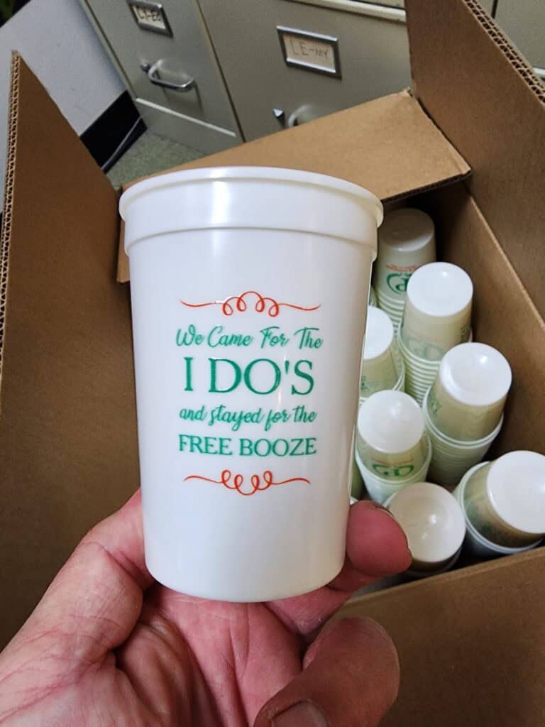 A hand holding a plastic cup that reads "We Came For The I DO'S and stayed for the FREE BOOZE" in colorful lettering. The background shows a box filled with similar cups and filing cabinets.