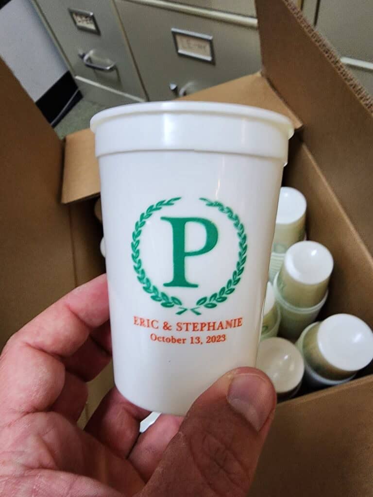 A hand holding a white cup featuring a green letter 'P' surrounded by a laurel wreath, with the text "Eric & Stephanie" and "October 13, 2023" printed in orange below it. The cup is held in front of an open cardboard box containing more cups, with filing cabinets visible in the background.