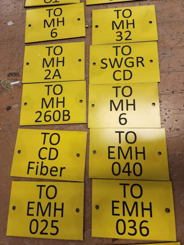 Yellow metal signage plates with black text containing various codes like 'TO MH 6', 'TO MH 2A', and 'TO EMH 025', laid out on a wooden surface.