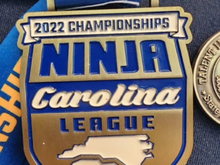 A large medal labeled '2022 Championships Ninja Carolina League' with a map outline of North and South Carolina on it. Surrounding it are other medals, partially visible, hanging on a surface, including one for 'Wood Ravin 2023' and another with partial text 'Duchen'. The medal features a combination of blue, white, and gold colors with a blue ribbon.
