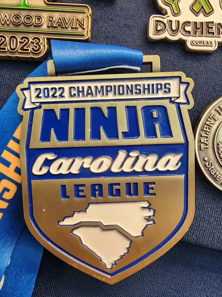 A large medal labeled '2022 Championships Ninja Carolina League' with a map outline of North and South Carolina on it. Surrounding it are other medals, partially visible, hanging on a surface, including one for 'Wood Ravin 2023' and another with partial text 'Duchen'. The medal features a combination of blue, white, and gold colors with a blue ribbon.