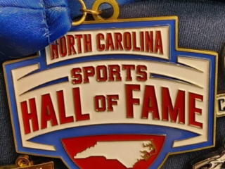 A collection of custom medals featuring the "North Carolina Sports Hall of Fame" medal prominently displayed in the center, with a blue ribbon attached. Other smaller medals surround it, each with unique designs and inscriptions, indicating various events or achievements.