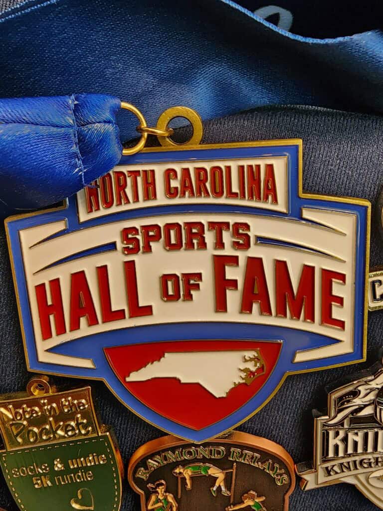 A collection of custom medals featuring the "North Carolina Sports Hall of Fame" medal prominently displayed in the center, with a blue ribbon attached. Other smaller medals surround it, each with unique designs and inscriptions, indicating various events or achievements.