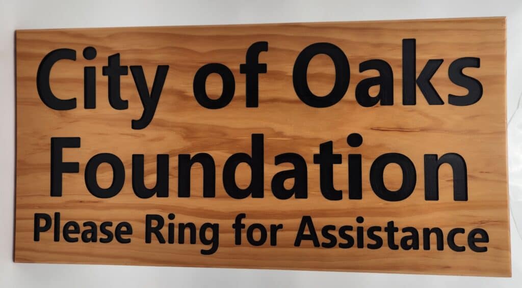 Wooden sign with black lettering that reads 'City of Oaks Foundation Please Ring for Assistance.'