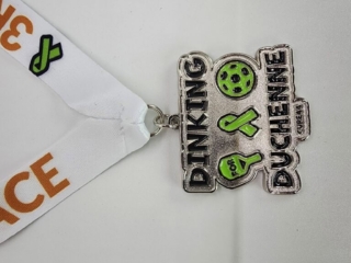 A custom silver medal with the text "Dinking for Duchenne" and illustrations of a pickleball paddle and ball, highlighted in green, attached to a white ribbon with orange and green design elements.