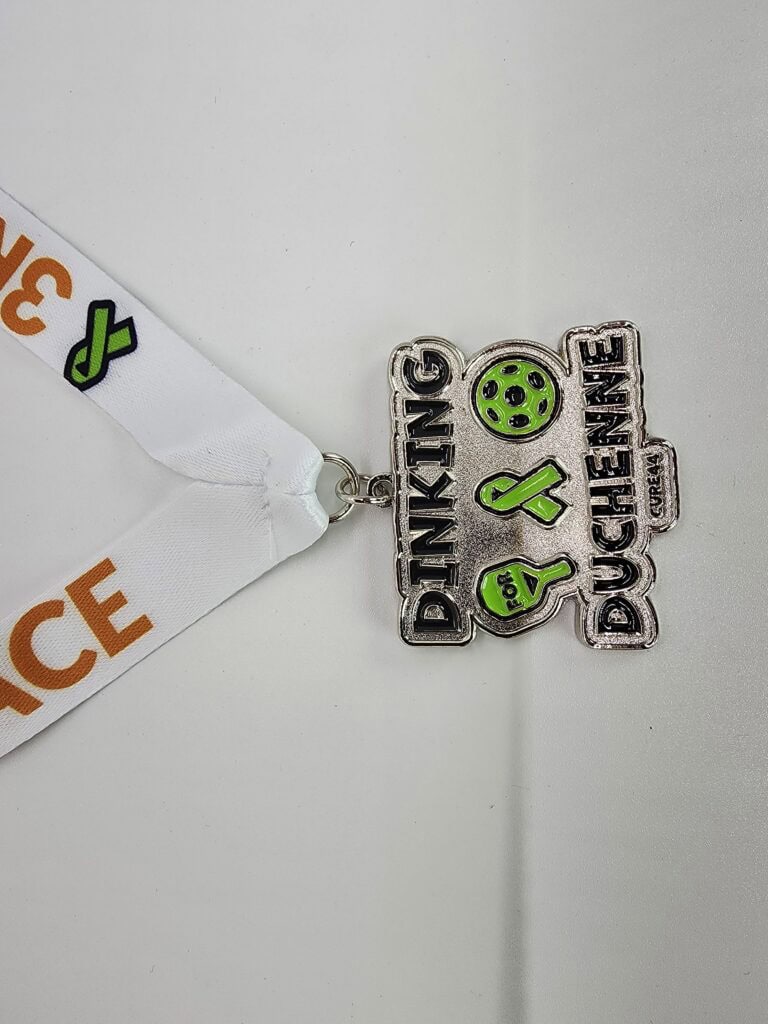 A custom silver medal with the text "Dinking for Duchenne" and illustrations of a pickleball paddle and ball, highlighted in green, attached to a white ribbon with orange and green design elements.