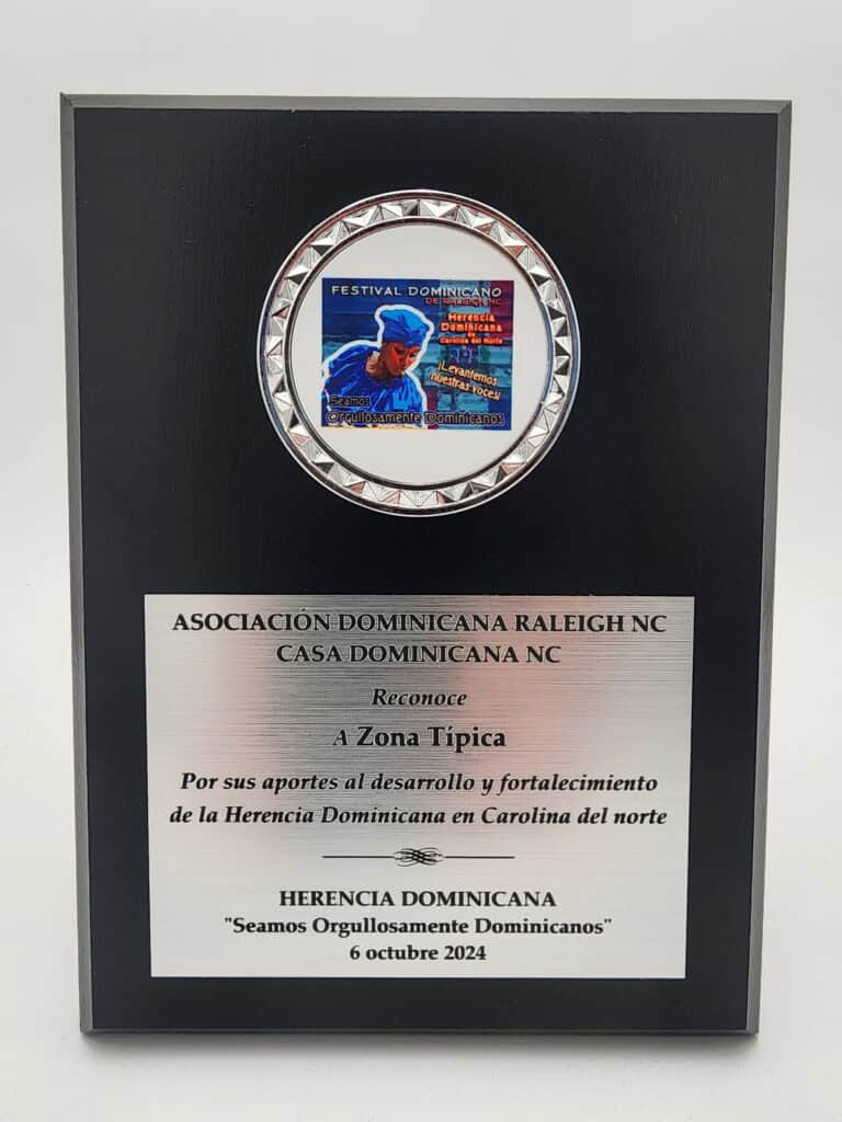 A black plaque with a silver trim features a colorful graphic at the top depicting a Dominican cultural festival. Below, engraved text recognizes the contribution of 'Zona Tipica' by the Dominican Association in Raleigh, NC, dated October 6, 2024.