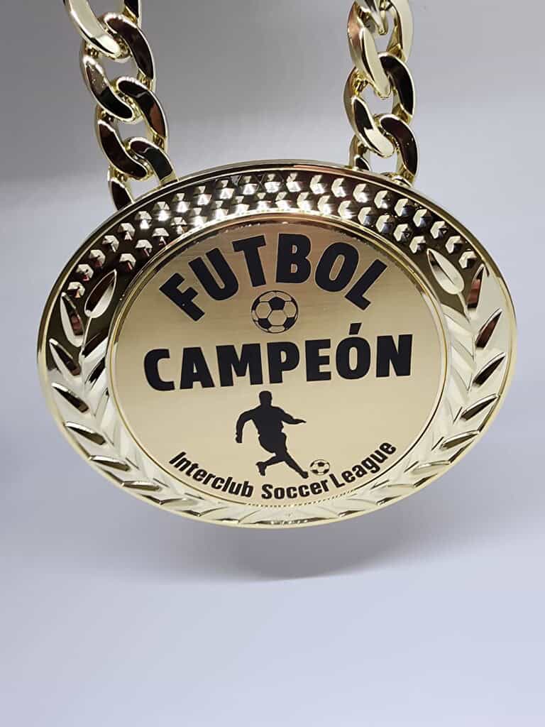 A gold medal with a chain, inscribed with 'FUTBOL CAMPE&Oacute;N' and 'Interclub Soccer League,' featuring a soccer player silhouette and a soccer ball design.