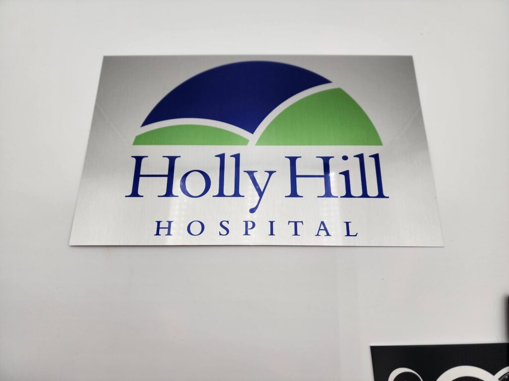 Sign with a logo featuring a blue and green semi-circle above the text "Holly Hill Hospital" on a grey background.
