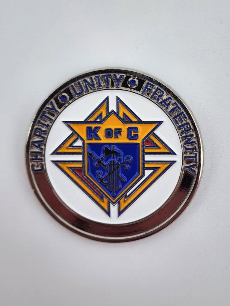 A commemorative coin featuring the Knights of Columbus emblem with the letters 'K of C' and symbols on a shield, surrounded by the words 'Charity', 'Unity', and 'Fraternity' around the edge.