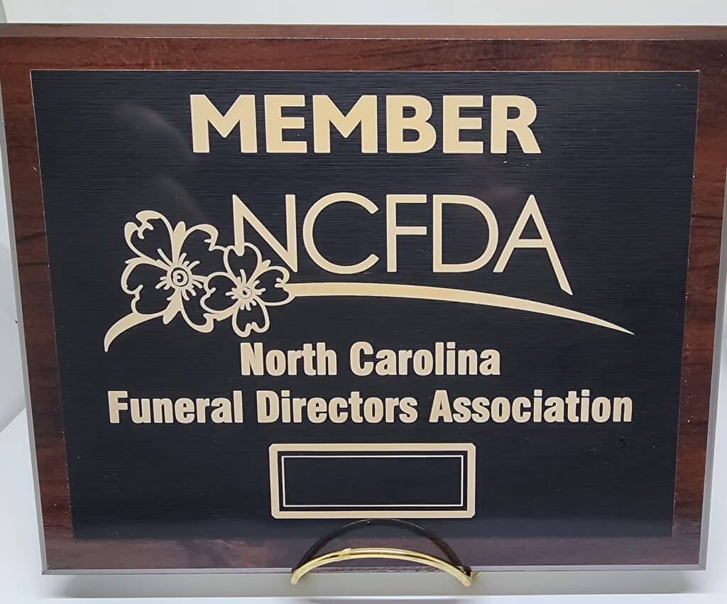 A wooden plaque with gold lettering displaying "MEMBER NCFDA North Carolina Funeral Directors Association" and floral embellishments.