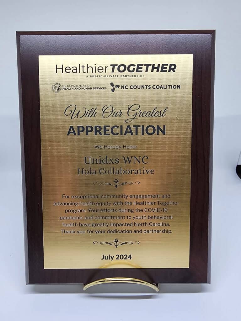 A wooden plaque with a gold plate, titled "Healthier Together." It expresses appreciation to Unidxs WNC Hola Collaborative for exceptional community engagement and advancing health equity, noting contributions during the COVID-19 pandemic. The plaque is awarded by the NC Department of Health and Human Services and NC Counts Coalition, dated July 2024.