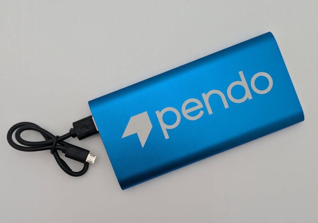 A blue portable power bank with the brand name "pendo" printed on it, connected to a black USB cable, placed on a white surface.