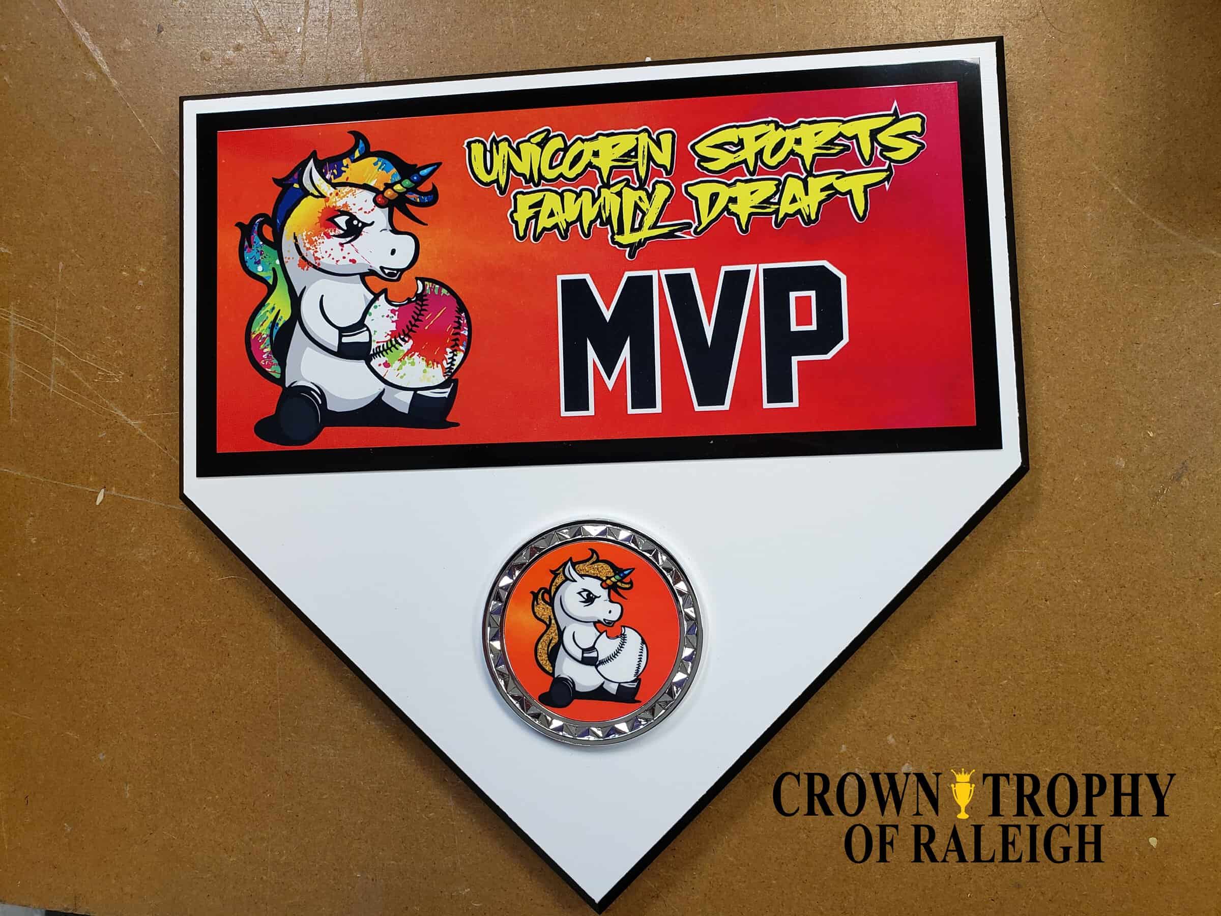 A recognition plaque shaped like a home plate, featuring a cartoon unicorn holding a rainbow-colored baseball with the text 'Unicorn Sports Family Draft MVP' on a red background. Below, there's a circular medallion with the same unicorn image. The plaque is branded with 'Crown Trophy of Raleigh.'