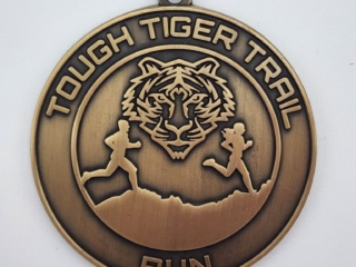 A bronze medal with the text 'Tough Tiger Trail Run' and a design featuring a tiger's face and two figures running along a trail.