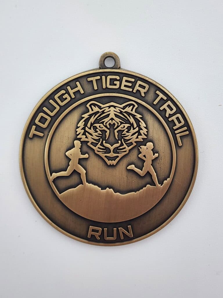 A bronze medal with the text 'Tough Tiger Trail Run' and a design featuring a tiger's face and two figures running along a trail.