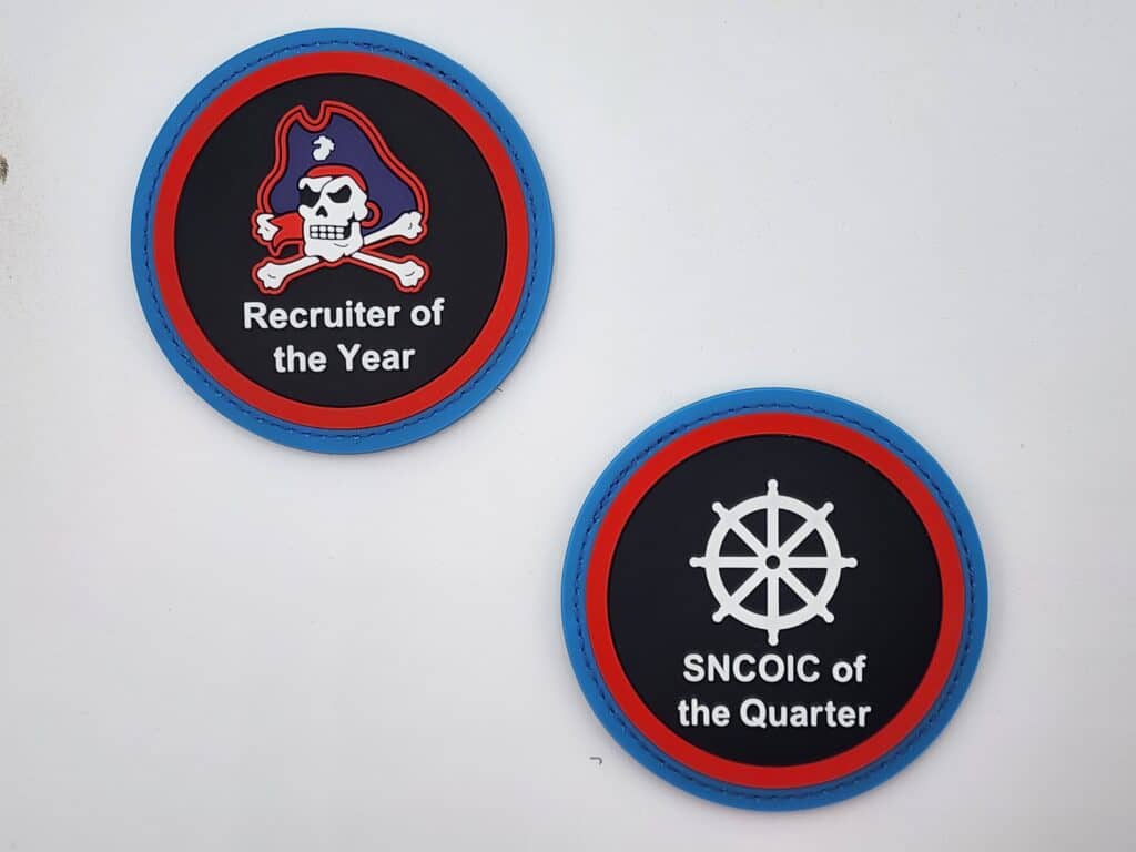 Two round patches on a white background. The left patch reads "Recruiter of the Year" with a pirate skull and crossbones design. The right patch reads "SNCOIC of the Quarter" with a ship's wheel design. Both have a black background with red and blue borders.